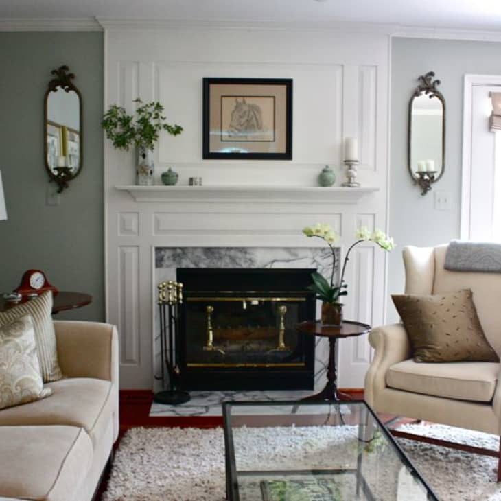 Living room with a white fireplace, beige sofa, wingback chair, glass coffee table, and decorative mirrors.