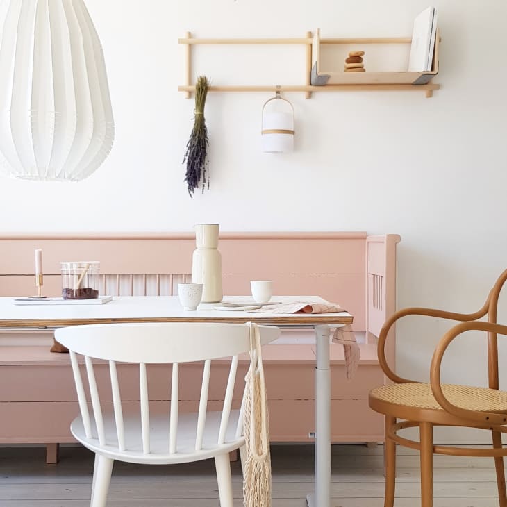Minimalist dining area with pink bench, white chairs, wooden table, and pendant light. Wall shelf with decor items.