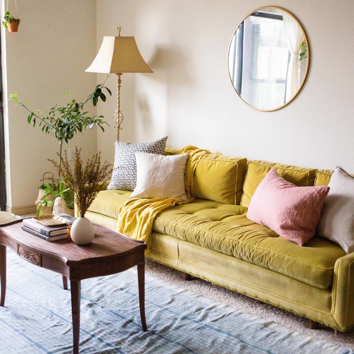 Yellow velvet sofa with pink and white pillows, wooden coffee table, potted plants, and a round mirror in a cozy living room.