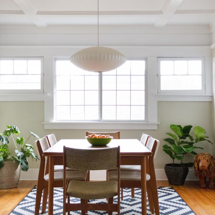 Dining room with wooden table, four chairs, geometric rug, pendant light, and potted plants by large windows.