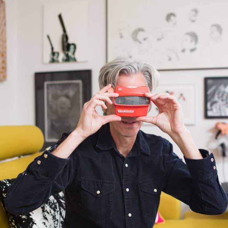 Person holding a red View-Master in a living room with yellow sofa and framed art on the wall.