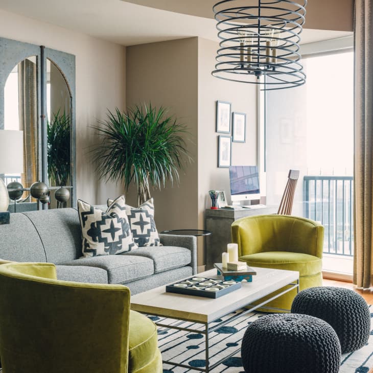 Modern living room with gray sofa, green chairs, geometric pillows, large plant, and a computer desk by the window.