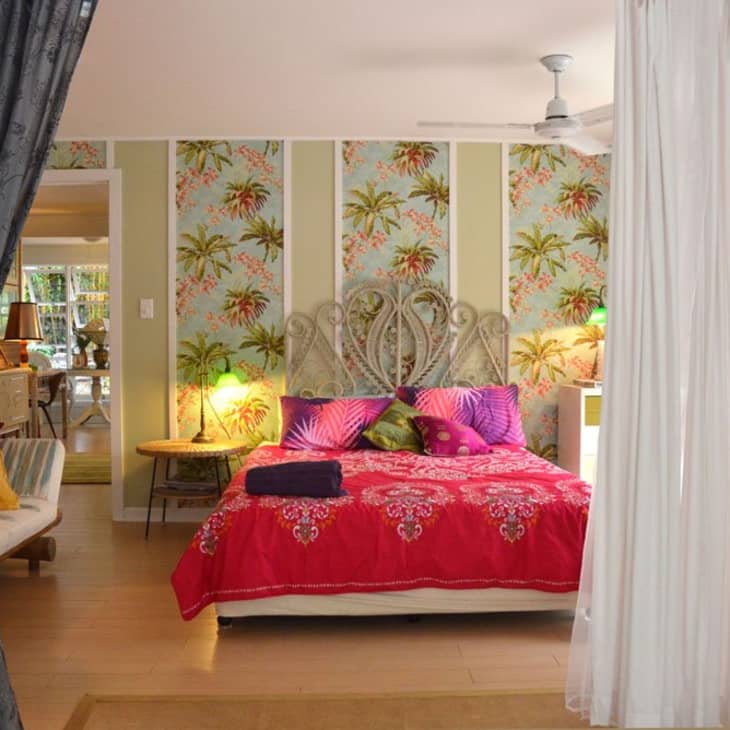 Bedroom with tropical wallpaper, ornate headboard, red bedspread, and colorful pillows, viewed through open curtains.