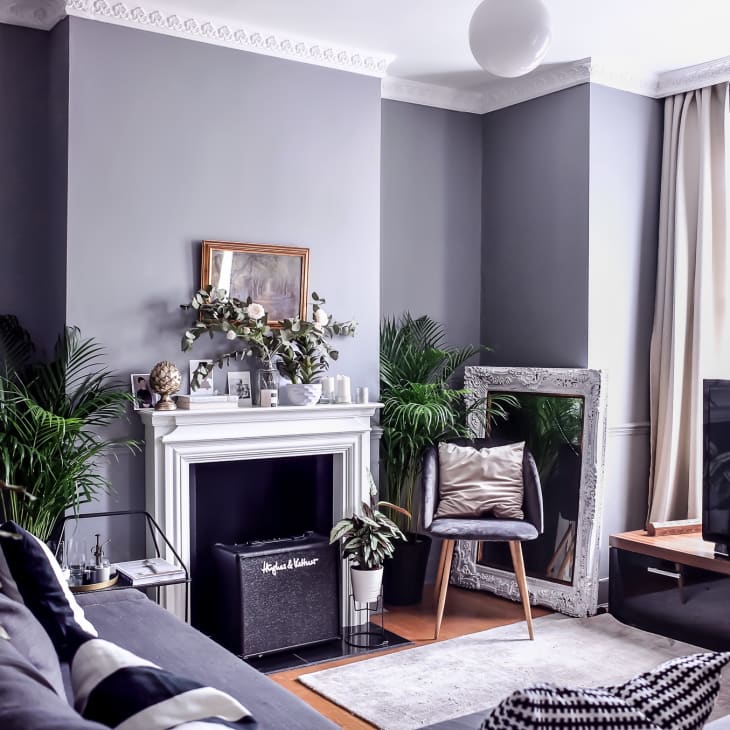 Cozy living room with gray walls, fireplace, plants, large mirror, TV, and a chair with a cushion.