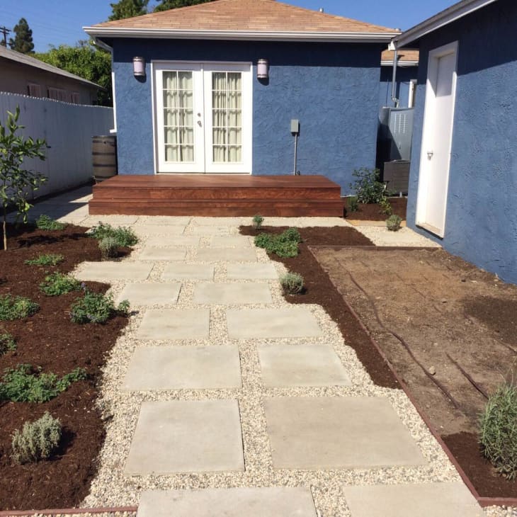 Stone path leading to blue house with French doors, surrounded by mulch and small plants.