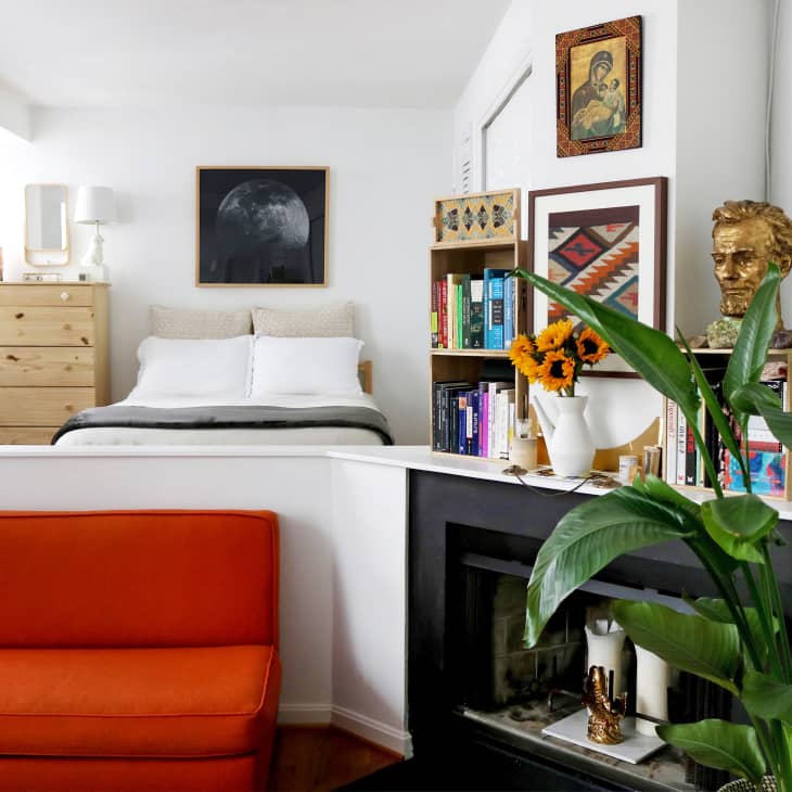 Bedroom with a white bed, orange sofa, bookshelf, sunflowers, and a gold bust on a mantel.