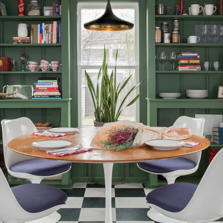 Dining room with green shelves, round wooden table, white chairs, and a plant centerpiece. Books and dishes on shelves.