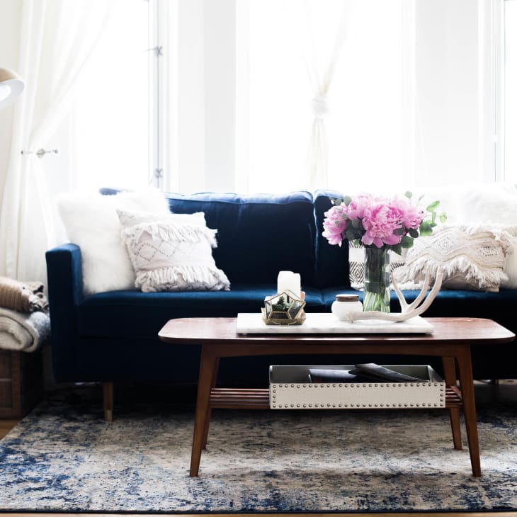 Living room with a navy sofa, pink flowers on a wooden coffee table, and a floor lamp beside stacked blankets.