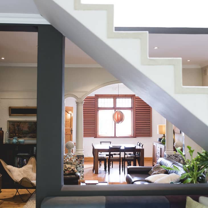 View of a dining room through a staircase, featuring a wooden table, chairs, and a large pendant light, with plants and decor.