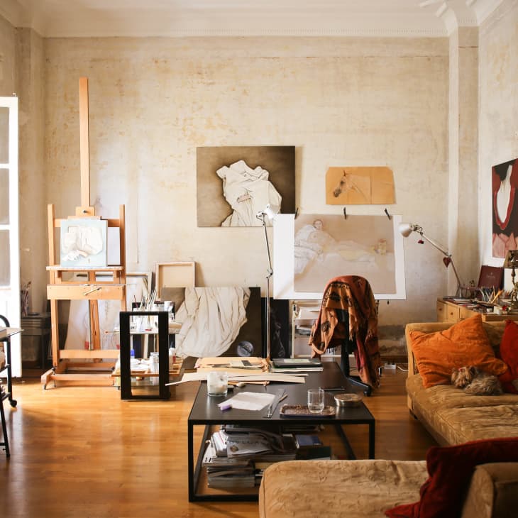 Art studio with easel, paintings, and a cozy sofa with cushions. Large windows let in natural light.