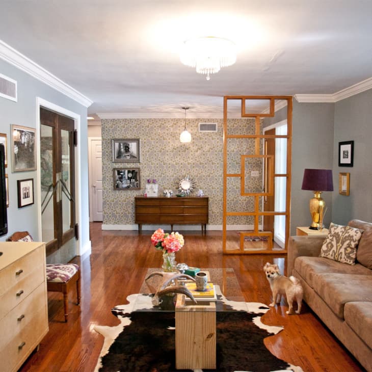Living room with beige sofa, TV, wooden floor, cowhide rug, and a small dog. Decor includes flowers and framed photos.