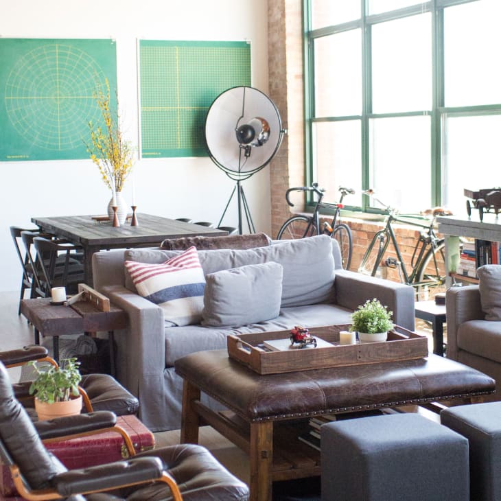 Loft living room with gray sofas, leather chairs, green wall art, and bicycles by large windows.
