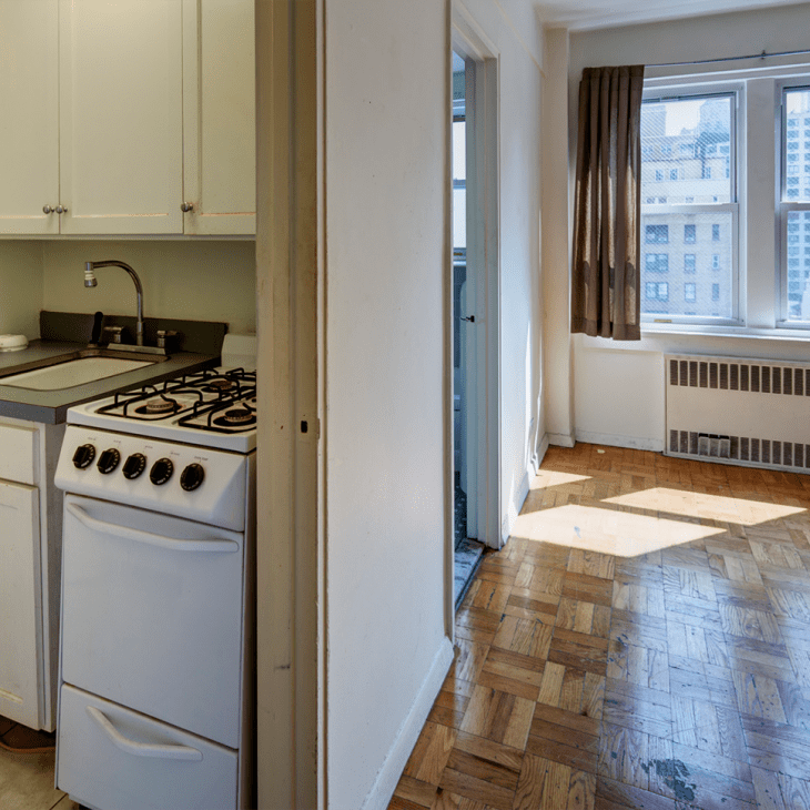 Small kitchen with white cabinets, gas stove, and sink, leading to a sunlit room with parquet flooring and large windows.