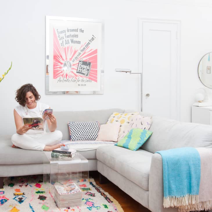 Woman reading on a light gray sectional sofa with colorful pillows, a glass coffee table, and a vibrant rug in a bright living room.
