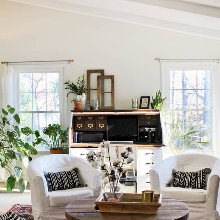 Bright living room with white armchairs, black patterned cushions, wooden desk, and various potted plants by large windows.
