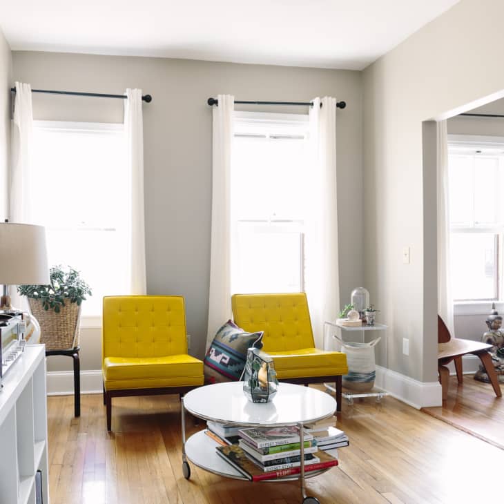 Living room with two yellow chairs, a round coffee table with books, and a potted plant near a window with white curtains.