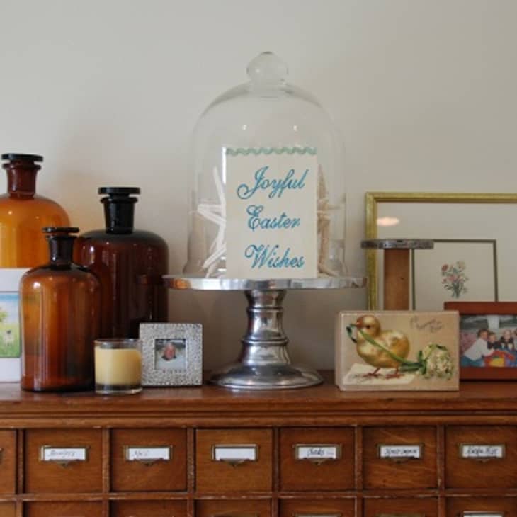 Vintage wooden cabinet with glass bottles, framed photos, and a cloche displaying "Joyful Easter Wishes."