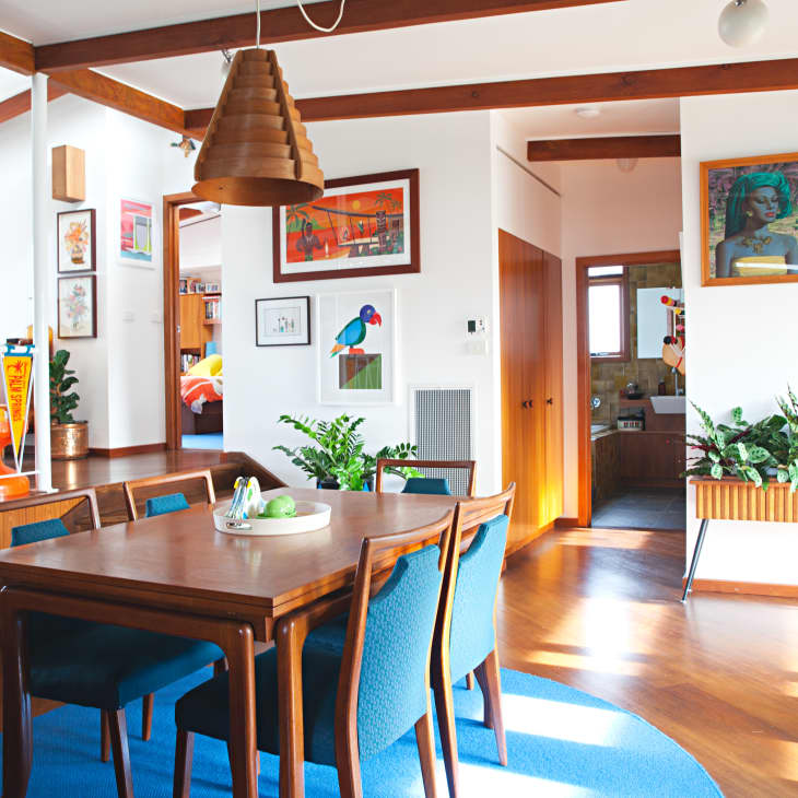 Mid-century modern dining room with wooden table, blue chairs, pendant light, and vibrant artwork on white walls.