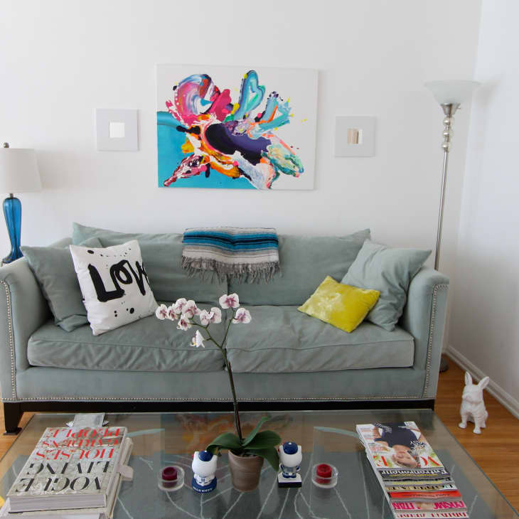 Light blue sofa with colorful pillows, abstract wall art, glass coffee table with magazines, and a white orchid in a vase.