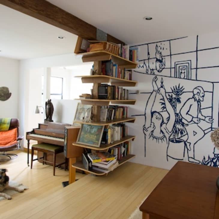 Living room with a mural wall, piano, bookshelf, brown sofa, and a dog lying on the floor.