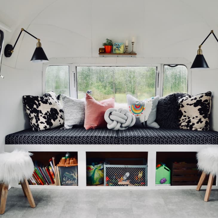 Camper interior with a cushioned bench, patterned pillows, wall sconces, and fluffy stools, overlooking a rainy landscape.