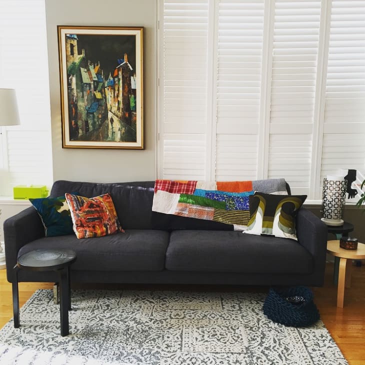 Dark gray sofa with colorful pillows and quilt, framed painting, and patterned rug in a living room with white shutters.
