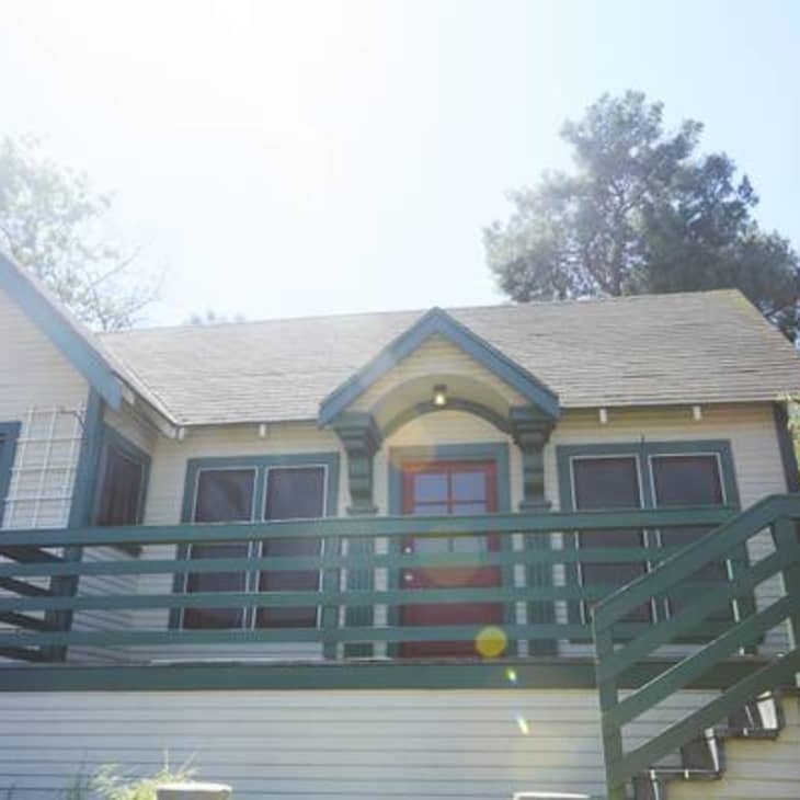 Charming house with green trim, red door, and a wooden deck, surrounded by trees and vibrant pink flowers.