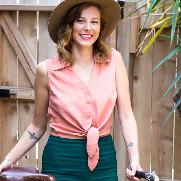Woman in a pink sleeveless top and green pants, wearing a hat, standing by a wooden fence with a bicycle.