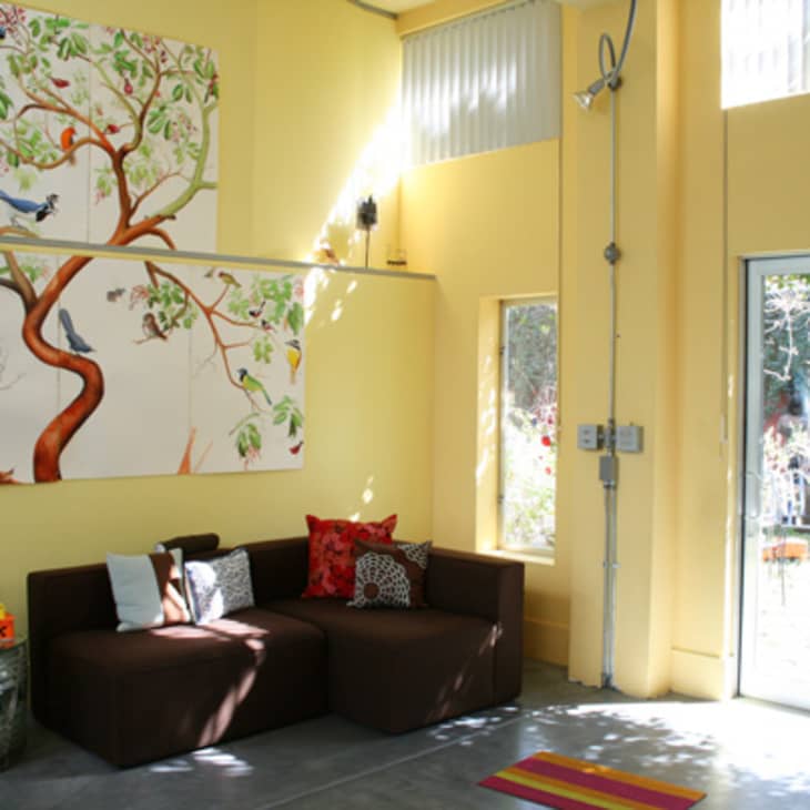 Bright living room with yellow walls, brown sofa, colorful cushions, large tree artwork, and glass door leading outside.