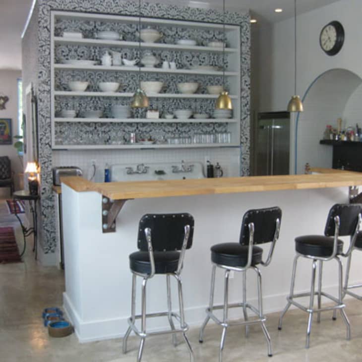 Home bar with wooden counter, four black bar stools, patterned shelves, and pendant lights in a modern kitchen setting.