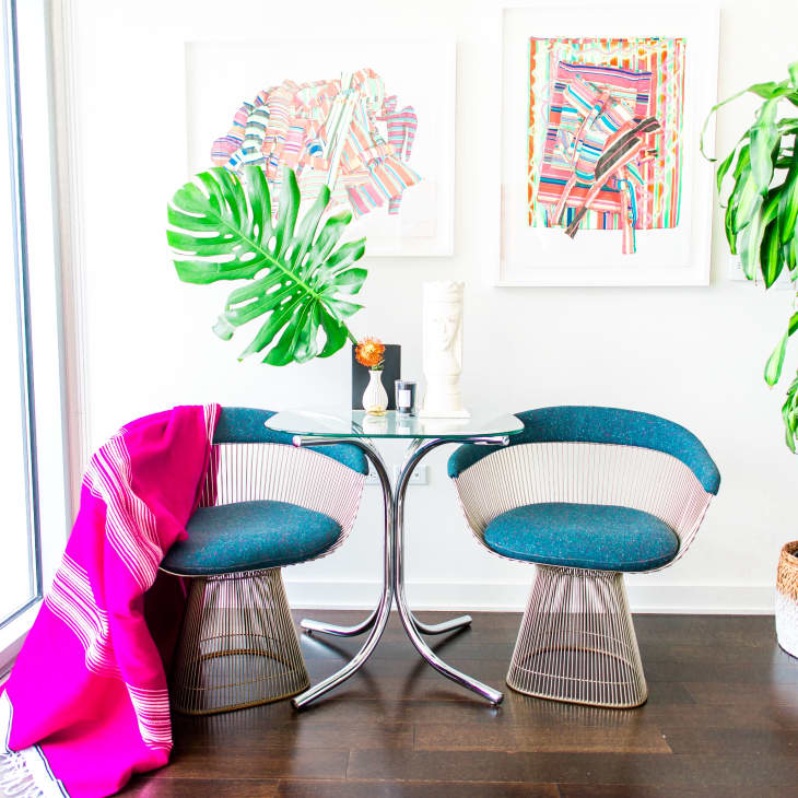 Two modern chairs with blue cushions and a glass table, pink throw, large potted plant, and colorful abstract art on the wall.