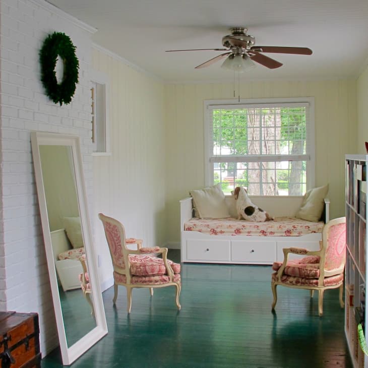 Sunroom with daybed, floral cushions, two vintage chairs, large mirror, ceiling fan, and potted plant by the window.