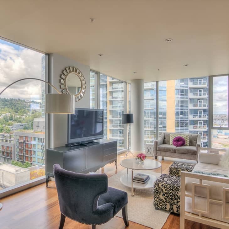 Modern living room with large windows, TV on a console, gray sofa, armchair, and decorative pillows.