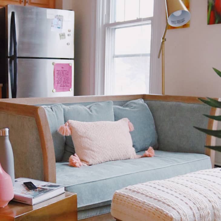 Cozy living room with a light blue sofa, pink pillow, knit blanket, and stainless steel kitchen appliances in the background.