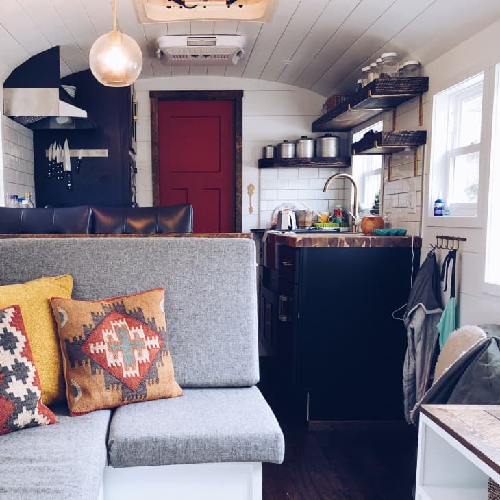 Tiny home interior with gray sofa, colorful patterned pillows, compact kitchen, red door, and open shelving.
