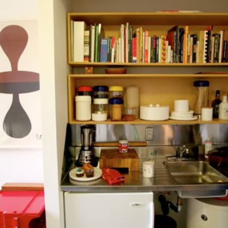 Small kitchen with open shelves, books, jars, and a man preparing food near a sink. Red storage unit and abstract art visible.