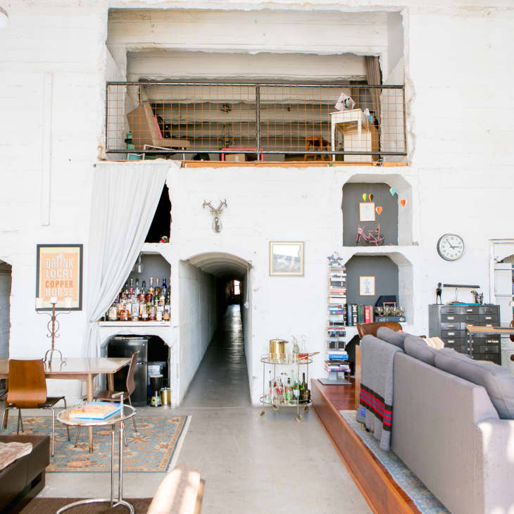 Loft-style living room with a gray sofa, bookshelves, bar cart, and dining table, featuring a mezzanine and eclectic decor.