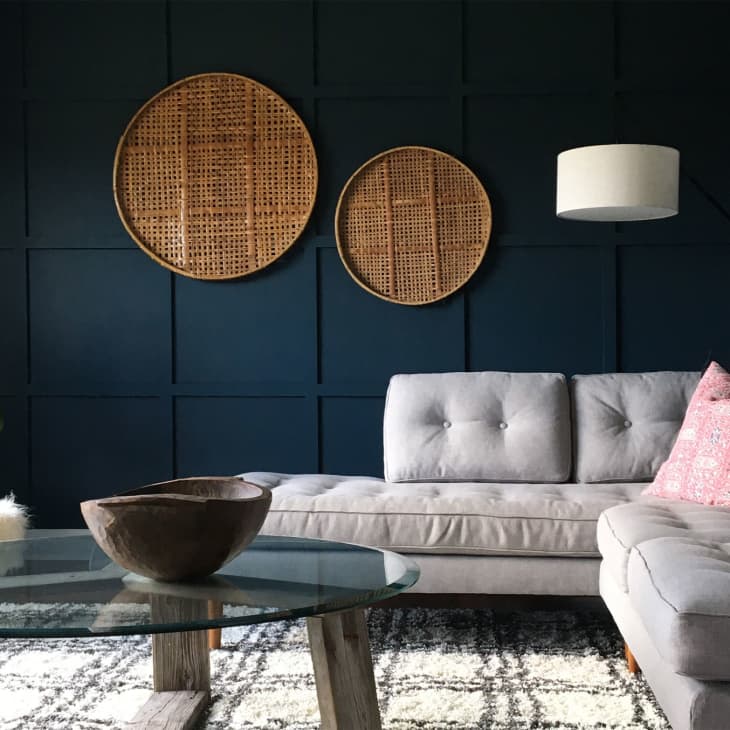 Living room with gray sectional sofa, glass coffee table, wicker wall decor, and a floor lamp against a dark paneled wall.