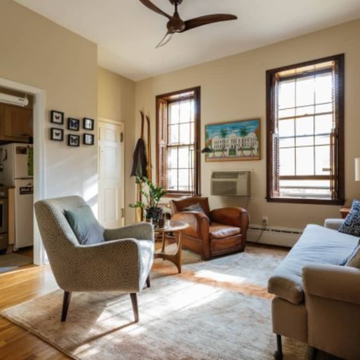 Cozy living room with armchairs, sofa, wall art, and large windows, adjacent to a small kitchen with vintage appliances.