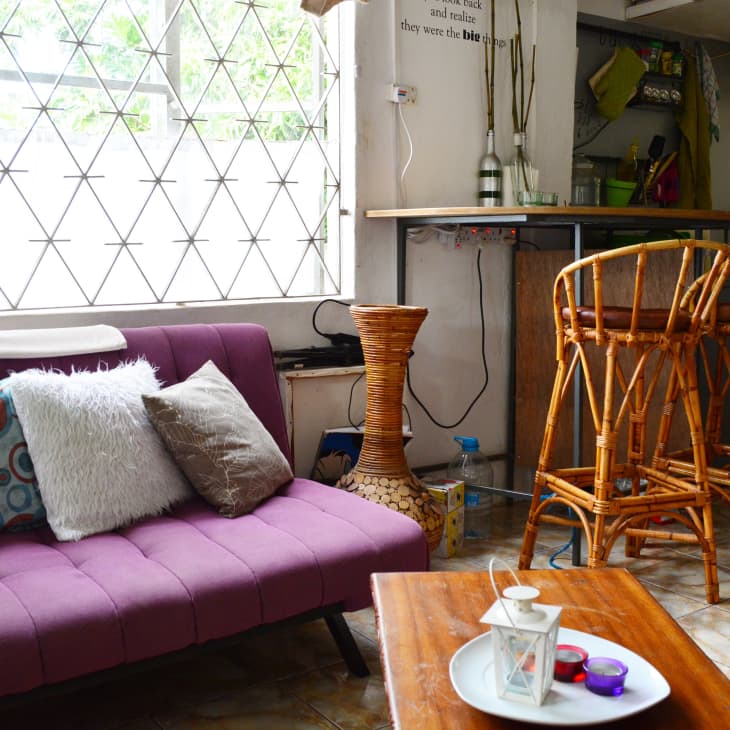 Purple sofa with patterned and fluffy pillows, wicker chairs, and a small bar area in a bright room with tiled floor.