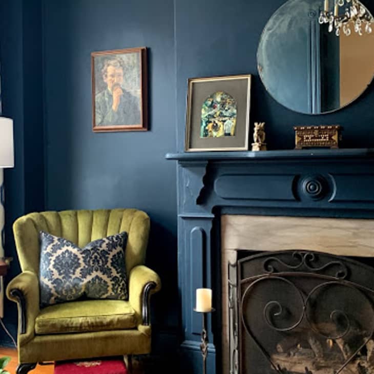 Dark blue living room with green armchair, patterned pillow, ornate fireplace, round mirror, and framed art.