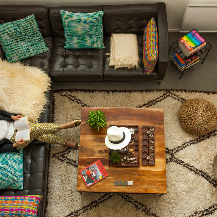 Cozy living room with a brown leather sectional, colorful pillows, wooden coffee table, and a person reading a book.