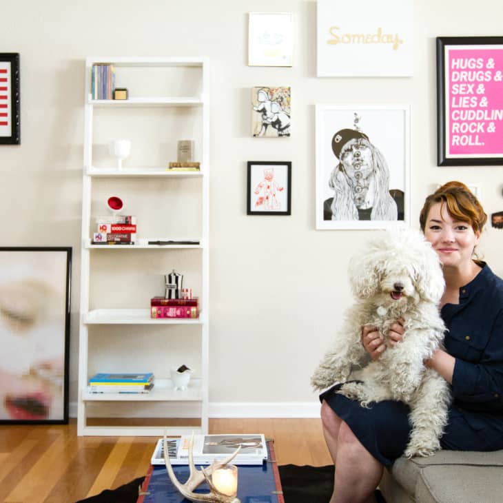 Woman sitting on a gray sofa holding a fluffy dog, surrounded by eclectic wall art and a white bookshelf.