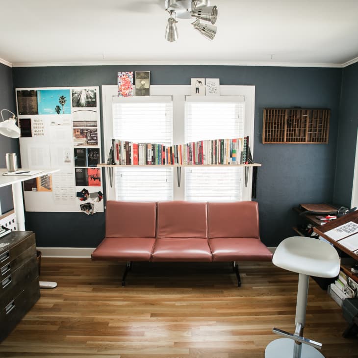 Home office with a brown leather sofa, wall art, books on a shelf, a drafting table, and a computer on a standing desk.