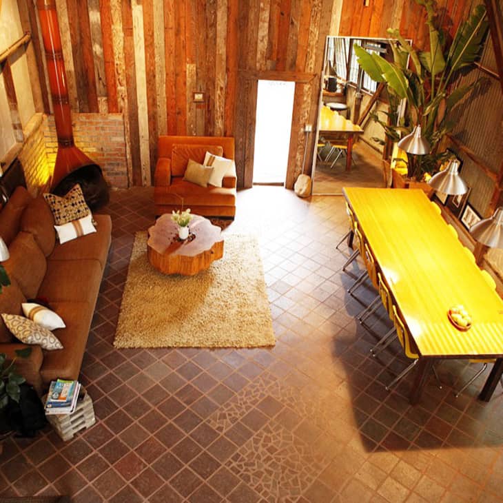 Rustic living and dining area with brown sofas, wooden table, large plants, and exposed brick and wood walls.
