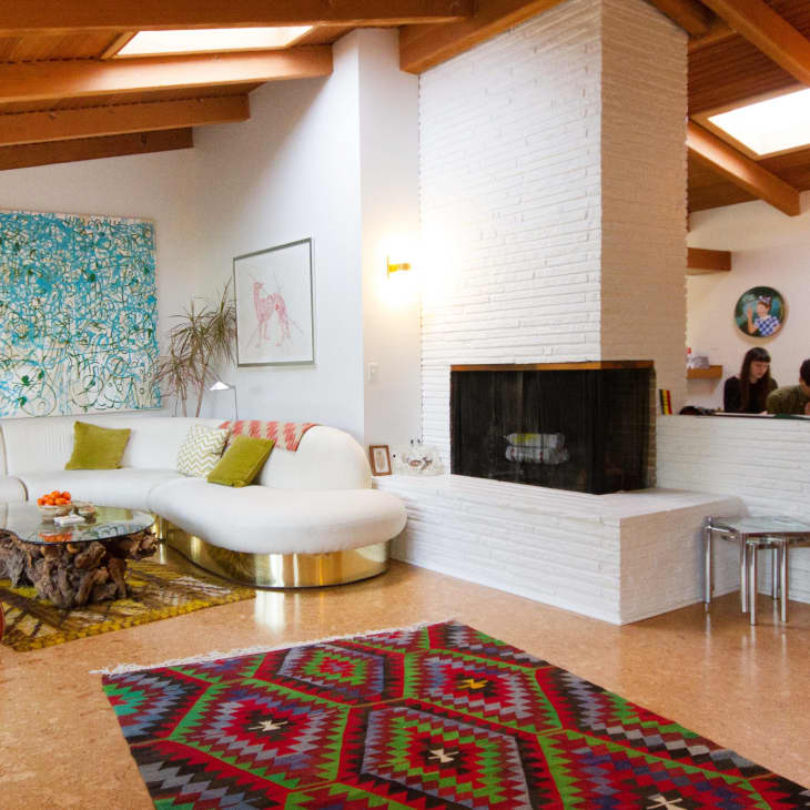 Mid-century modern living room with white sectional sofa, colorful abstract art, geometric rug, and central brick fireplace.