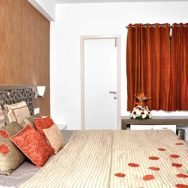 Bedroom with patterned headboard, beige and red pillows, orange curtains, and a green chair at a desk.