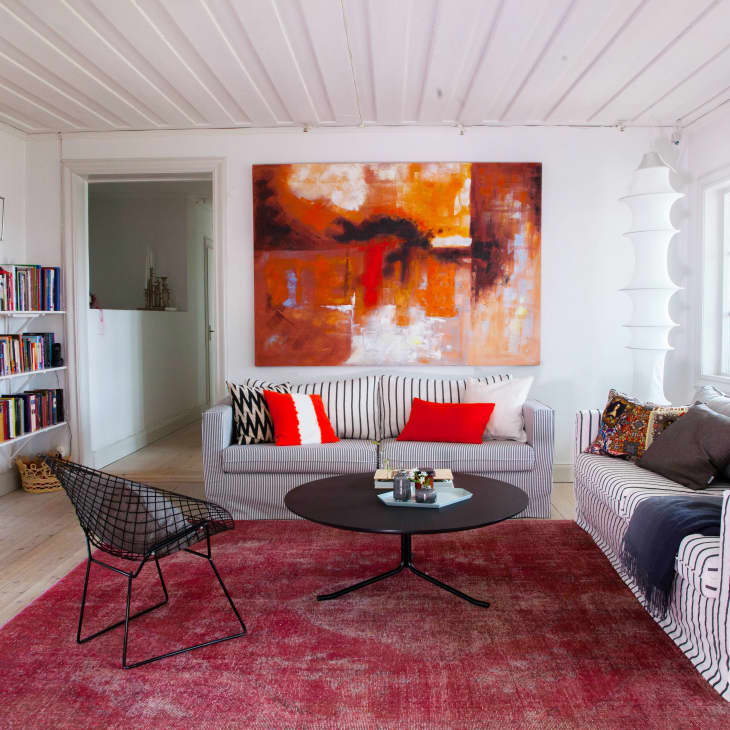 Living room with striped sofas, red cushions, abstract painting, bookshelves, and a black wire chair on a red rug.