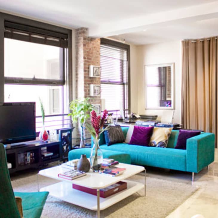 Modern living room with teal sofa, colorful cushions, TV, and coffee table with books and flowers.