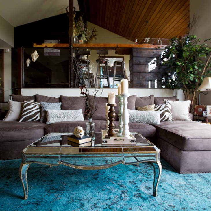 Brown sectional sofa with patterned pillows, mirrored coffee table, candles, and a large plant in a cozy living room.
